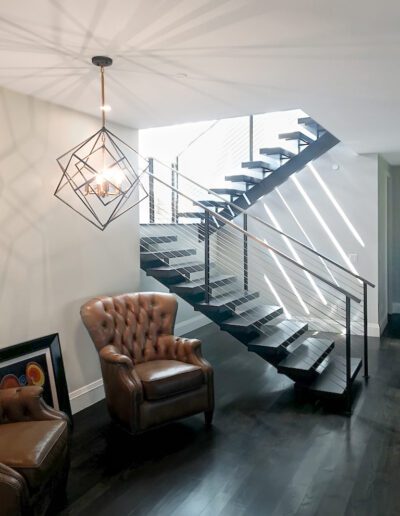 A modern interior featuring a staircase with black railings and floating wooden steps, an elegant geometric pendant light, and a classic leather armchair.