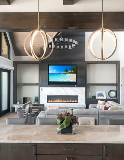 A modern living room with a fireplace, large tv, and circular chandeliers, viewed over a kitchen island.