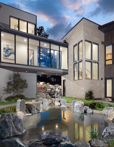 Modern two-story house with large windows at dusk featuring a landscaped garden with a pond and rocks.