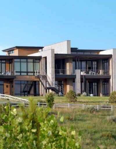 Modern residential building with large glass windows and a mix of wooden and concrete elements, situated in a green landscape.