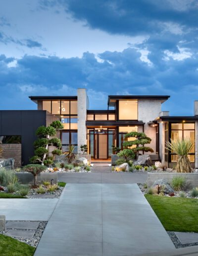 Modern luxury house with illuminated windows at twilight, featuring a landscaped garden and concrete driveway.