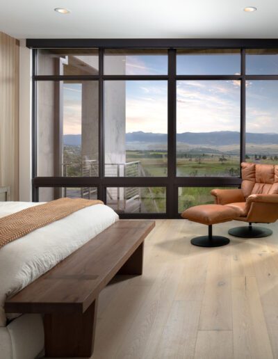 Modern bedroom with a panoramic view of the countryside through large windows, featuring a minimalist design and earthy tones.