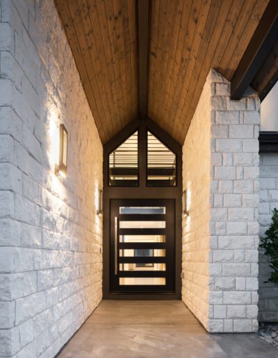 An illuminated modern entrance with a wooden ceiling, stone walls, and a stylish black door with horizontal windows.